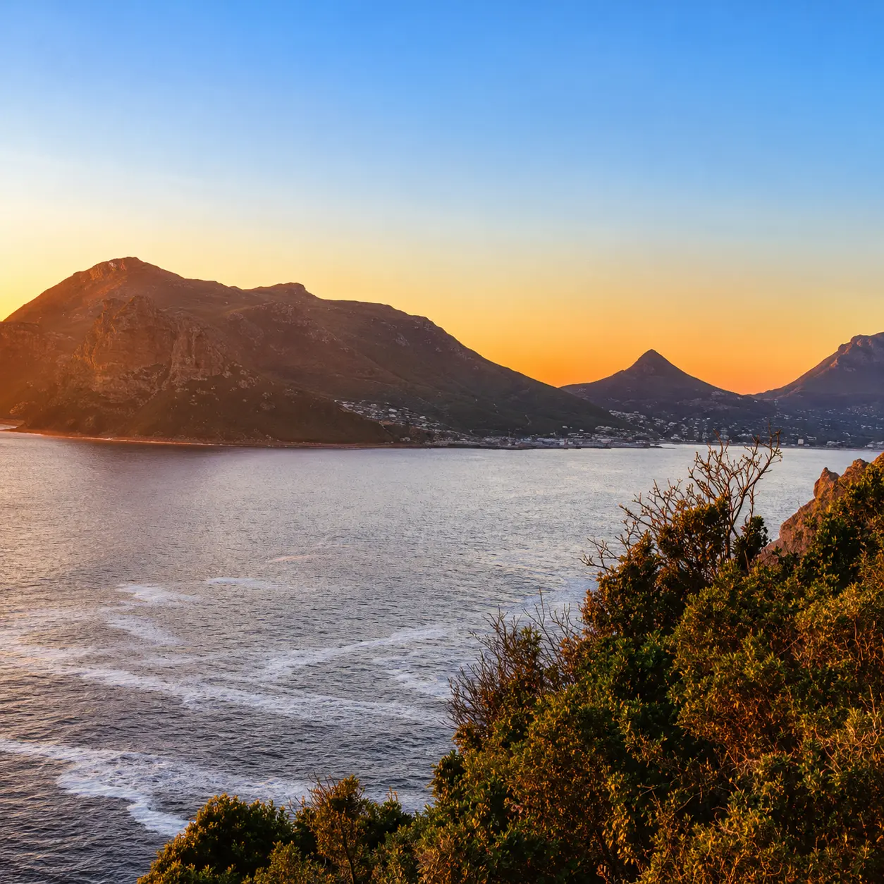 Golden sunrise over Hout Bay with calm ocean water, rugged mountains and coastal vegetation in Cape Town.
