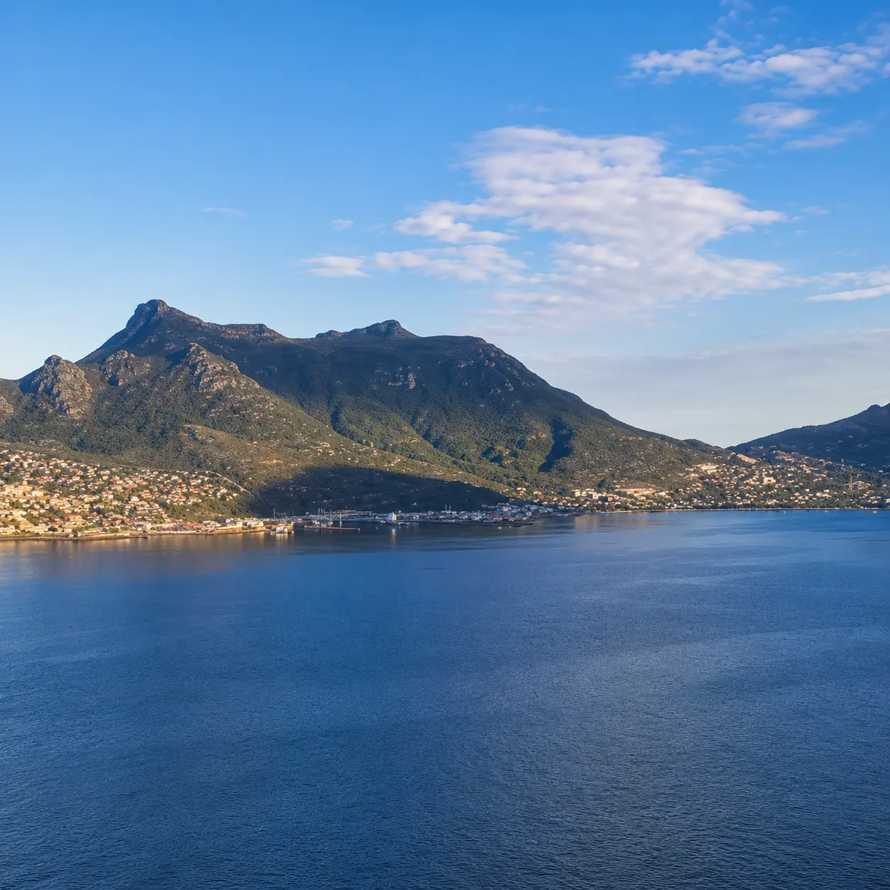 Panoramic view across Hout Bay toward the coastal town and surrounding mountains under a clear blue sky in Cape Town.