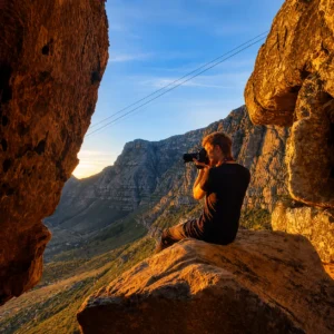 Photographer sitting on a rocky ledge and photographing Table Mountain cliffs in warm golden morning light in Cape Town
