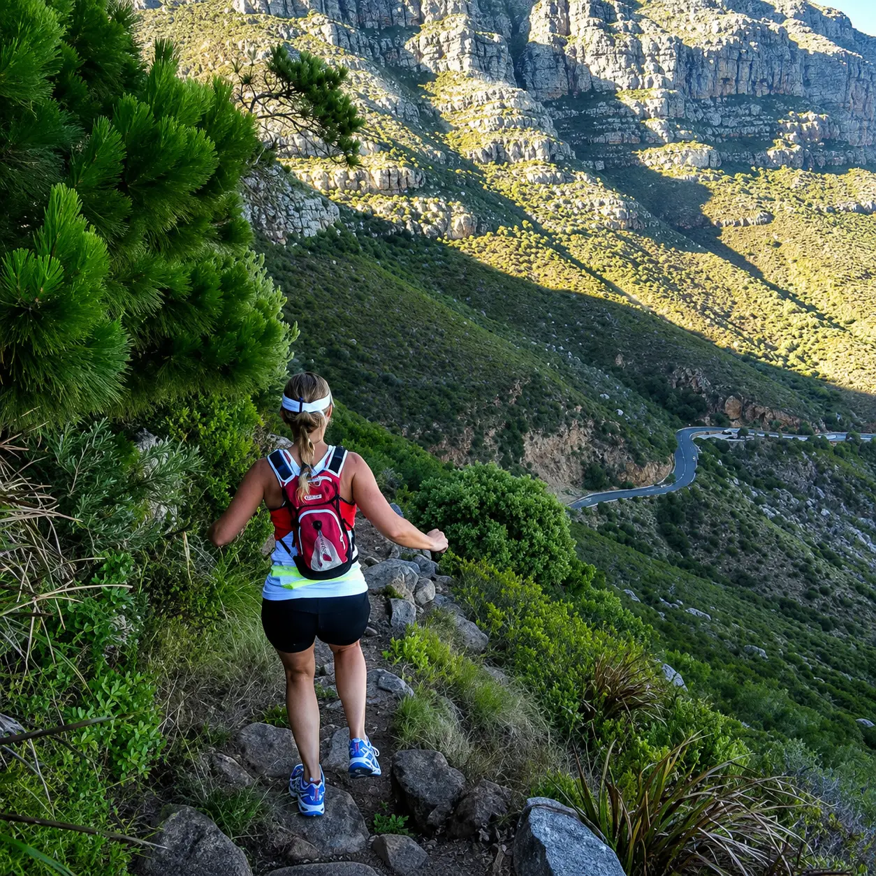 Trail runner descending a rugged mountain path through dense vegetation with dramatic rocky cliffs and a winding road below.