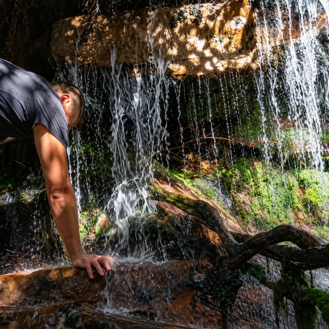 Hiker beside a small mountain waterfall cascading over rocks and tree roots along a scenic Cape Town hiking trail.