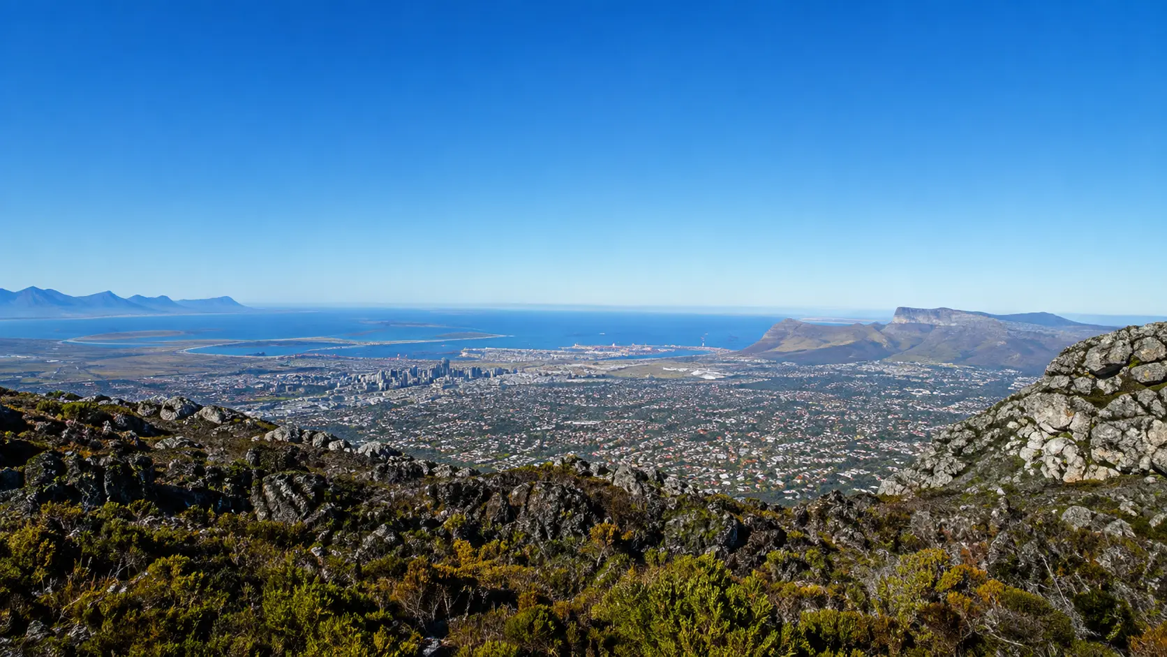 Panoramic mountain view overlooking Cape Town, Table Bay, the city centre and distant mountain ranges under a clear blue sky.