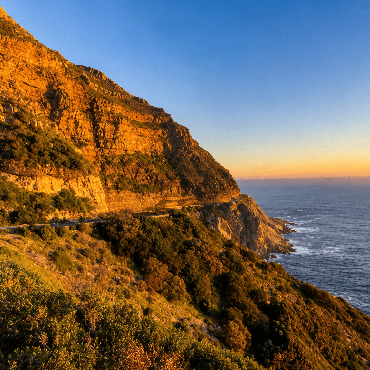 Rugged coastal cliffs glowing in golden-hour sunlight above a winding road and the Atlantic Ocean near Cape Town.