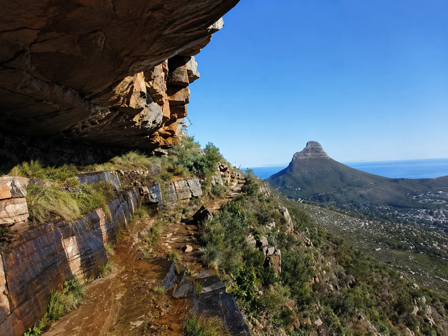 Hiking trail beneath a rocky overhang on Table Mountain with Lion’s Head and the Cape Town coastline in the background.