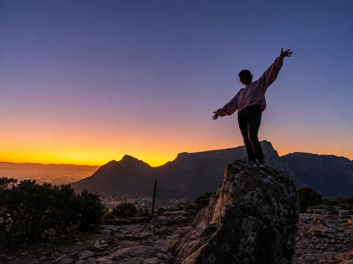 Hiker standing with arms outstretched on a rocky summit at sunrise, overlooking Cape Town and Table Mountain.